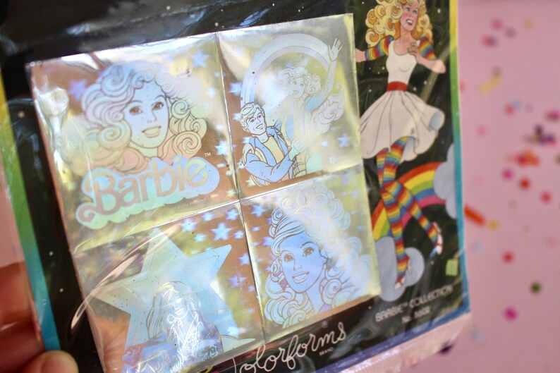 Puede incluir: Un paquete de pegatinas hologr&aacute;ficas con im&aacute;genes de Barbie y Ken. Las pegatinas est&aacute;n en un paquete de pl&aacute;stico transparente con fondo negro. El paquete tiene el texto "Barbie" y "Colorforms" impreso en &eacute;l.