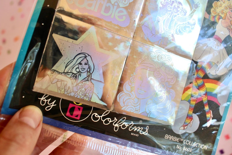 Puede incluir: Un paquete de pegatinas hologr&aacute;ficas con im&aacute;genes de mu&ntilde;ecas Barbie. Las pegatinas son de la marca Colorforms y forman parte de la colecci&oacute;n Barbie. El n&uacute;mero del paquete es 8602.