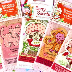 May include: Five Strawberry Shortcake themed car air fresheners. Each air freshener is a different scent and features a different character. The scents are bubblegum, strawberry, banana, and two strawberry scents. The characters are Strawberry Shortcake, a cat, a monkey, and two more Strawberry Shortcake characters.