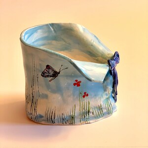 Może przedstawiać: Jasnoniebieska ceramiczna doniczka z namalowanym motylem i czerwonymi kwiatami. Doniczka ma podwinięty brzeg i niebieską wstążkę zawiązaną wokół górnej części.