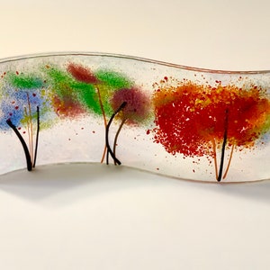 Op de afbeelding: Een kunstwerk van glas met drie gestileerde bomen met kleurrijk, gevlekt gebladerte. De bomen staan ​​op een witte achtergrond en zijn gemonteerd op een gebogen glazen voet.