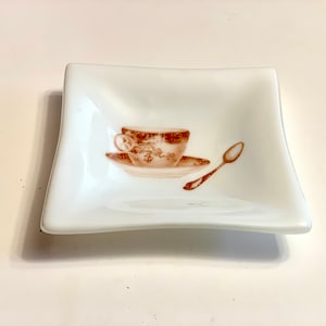 May include: A white ceramic dish with a brown image of a teacup and saucer with a spoon.