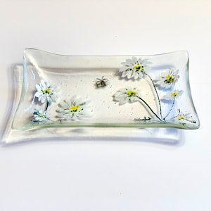 Może przedstawiać: Przezroczysta szklana miska z wzorem białych stokrotek. Miska ma lekko zakrzywiony kształt i idealnie nadaje się do przechowywania drobnych przedmiotów.