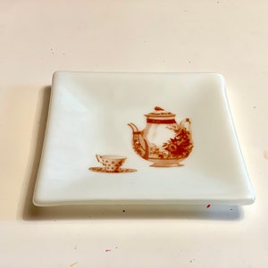 May include: A white square ceramic dish with a brown image of a teapot and a teacup. The teapot and teacup are decorated with a floral pattern.