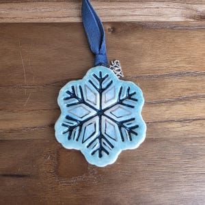 May include: A ceramic snowflake ornament with a light blue glaze and silver accents. The snowflake design has a dark blue outline and is attached to a blue ribbon. A small silver charm with the number "2023" is attached to the ribbon.