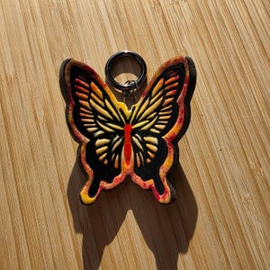 Puede incluir: Un llavero con forma de mariposa con un anillo de metal. La mariposa tiene un contorno negro con detalles amarillos, naranjas y rojos. El llavero está sobre una superficie de madera.