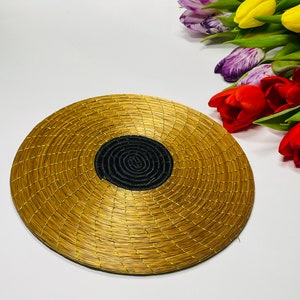Puede incluir: Un plato redondo tejido de color dorado con un centro negro. El plato está hecho de materiales naturales y tiene un aspecto rústico y hecho a mano.