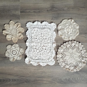 May include: A collection of five off-white and beige crocheted doilies in various shapes and sizes. The set includes one rectangular piece with a floral motif, two circular doilies, and two smaller flower-shaped pieces with intricate openwork.