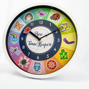 May include: A round wall clock with a white frame and a dark blue center featuring the text Bee Time Keeper. The clock face has a colorful radial background with illustrations of a hedgehog, bird, ladybug, bee, caterpillar, turtle, flower, owl, spider, dragonfly, rainbow, and butterfly.