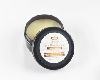 May include: A black jar with a white label that reads "SHCO Spa at Home Company Citrus Sunshine Sugar Scrub". The ingredients are listed on the label: Sugar, Apricot Oil, Aloe Vera Gel, Corn Starch, Essential Oils. The jar is open and the scrub is visible inside.