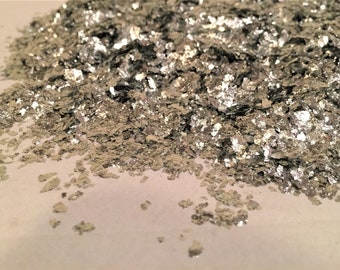May include: A pile of silver-colored, irregularly shaped flakes. The flakes have a metallic sheen and reflect light, creating a shimmering effect. The material appears to be scattered across a light-colored surface.