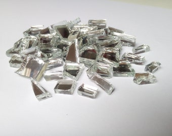 May include: A pile of irregularly shaped, reflective, silver-colored glass pieces. The pieces vary in size and shape, with some appearing rectangular and others more triangular. The glass has a shiny, mirrored surface.