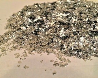 May include: A pile of silver-colored, irregularly shaped flakes. The flakes have a metallic sheen and reflect light, creating a shimmering effect. The material appears to be loose and scattered.