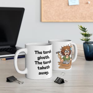 Può includere: Una tazza in ceramica bianca con un'illustrazione a cartone animato di una persona con capelli spettinati e occhiali protettivi, che tiene un martello e un cannello sopra un banco da lavoro, con il testo "The torch giveth. The torch taketh away.".