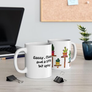 May include: A white ceramic mug featuring a colorful illustration of various cacti in pots on wooden shelves and the black text "Sassy, Classy and a little bit spiky" printed on the side. The mug has a standard handle and a smooth finish.