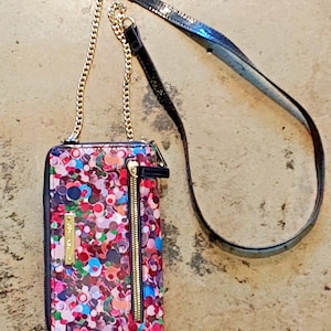 May include: A wallet-style crossbody bag with a gold chain and a long, black leather strap. The bag has a vibrant pattern of multicoloured circles and a gold zip. A small gold-coloured metal tag is on the front.