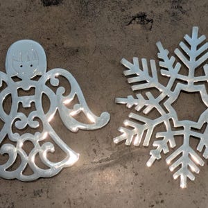 Puede incluir: Dos adornos navideños plateados: un ángel y un copo de nieve. El ángel tiene un diseño calado con alas y un halo. El copo de nieve tiene un diseño simétrico de seis puntas. Ambos están sobre una superficie marrón texturizada.