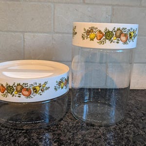 May include: Two clear glass storage jars with white lids and rims. The rims feature a colourful pattern of fruits and vegetables. One jar is short and wide, the other is tall and cylindrical.