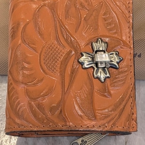 Op de afbeelding: Oranje leren portemonnee met een reliëf bloemenpatroon en een zilverkleurige kruisvormige sluiting. De portemonnee heeft een ritstrekker en een gevouwen ontwerp. Het leer heeft een verweerde uitstraling.