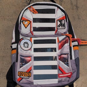Puede incluir: Mochila con diseño temático espacial en gris, blanco, naranja y negro. Cuenta con múltiples parches, incluyendo los textos "MARS AVIATOR" y "SPRAYGROUND". Dispone de asa superior, correas ajustables y varios compartimentos con cremallera.