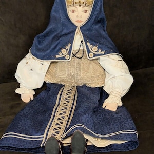 Puede incluir: Muñeca hecha a mano vestida con atuendo tradicional. La muñeca presenta una falda y capa azules estampadas, una diadema dorada y un corpiño color crema. La cara de la muñeca está pintada con rasgos delicados y lleva zapatos negros.