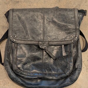 Peut inclure: Un sac à dos en cuir gris anthracite foncé avec une finition vieillie, doté d'un rabat avec des clous métalliques, d'une poche avant avec une tirette de fermeture éclair et du texte "the sak" imprimé sur une petite languette centrale.