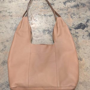 May include: A large, light brown leather shoulder bag with a rounded shape. The bag has a single, long strap in a darker brown color. The bag appears to be empty and has a simple, minimalist design.