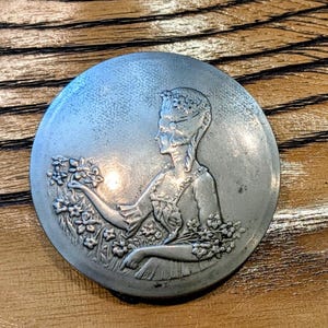 Könnte beinhalten: Runde, silberfarbene Metallmedaille mit dem Profil einer Frau, umgeben von Blumen. Die Frau trägt einen Blumenkranz und hält Blumen. Die Medaille liegt auf einem Holzgrund.