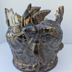 May include: A ceramic planter shaped like a group of sea turtles. The planter is black with a metallic sheen and has a gold-colored glaze. The turtles are stacked on top of each other, with the largest turtle at the bottom and the smallest turtle at the top.