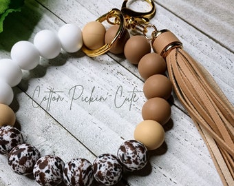 May include: A brown, white and black silicone beaded keyring with a gold key ring and a brown leather tassel. The keyring has the text "Cotton Pickin' Cute" on it.