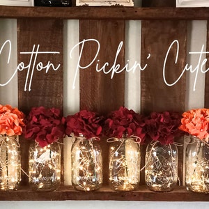 May include: Six mason jars with string lights inside, decorated with fall-colored flowers. The jars are arranged on a wooden shelf with the text "Cotton Pickin' Cute" above them.