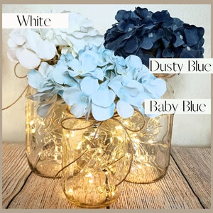May include: Three mason jars with fairy lights inside, decorated with white, baby blue, and dusty blue hydrangea flowers. The jars are tied with twine.