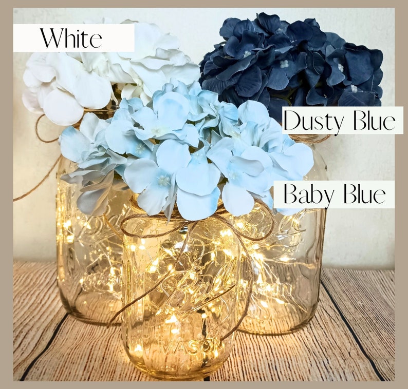 May include: Three mason jars with fairy lights inside, decorated with white, baby blue, and dusty blue hydrangea flowers. The jars are tied with twine.