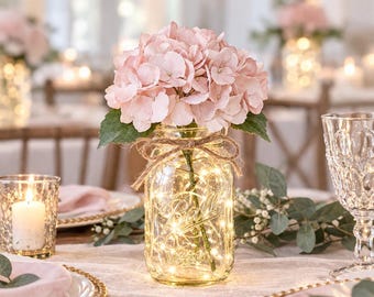 May include: A clear glass Ball mason jar filled with warm white fairy lights and topped with a bouquet of light pink hydrangea flowers. The jar is tied with a natural jute twine bow around the neck, creating a decorative centrepiece.