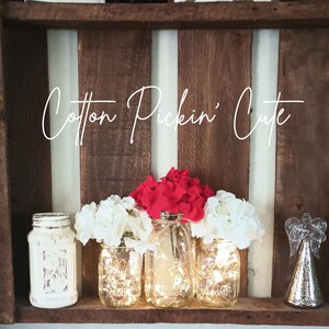 May include: Three mason jars with fairy lights inside, decorated with white and red flowers. The jars are sitting on a wooden shelf with the text "Cotton Pickin' Cute" above them.