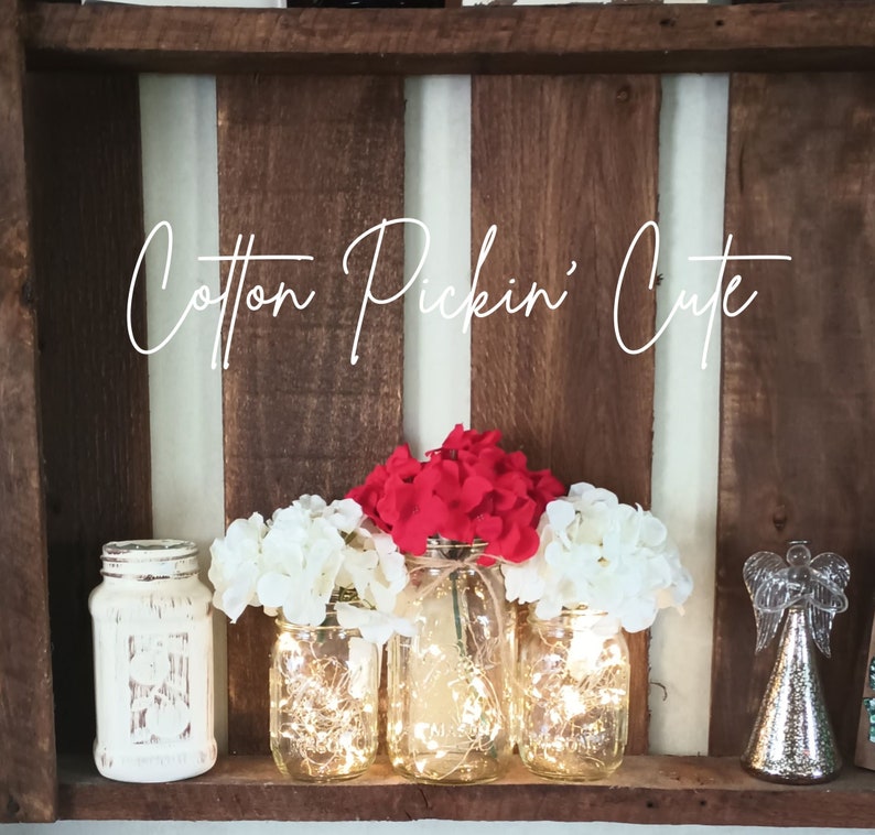 May include: Three mason jars with fairy lights inside, decorated with white and red flowers. The jars are sitting on a wooden shelf with the text "Cotton Pickin' Cute" above them.