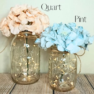 May include: Two mason jars with string lights inside, decorated with pink and blue hydrangeas. The larger jar is labeled "Quart" and the smaller jar is labeled "Pint".