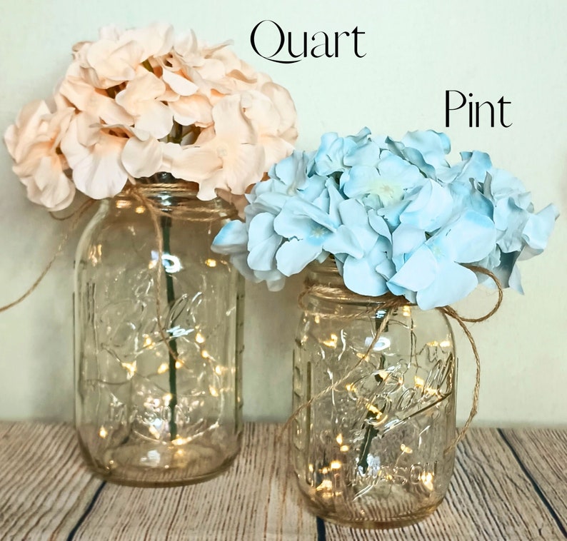 May include: Two mason jars with string lights inside, decorated with pink and blue hydrangeas. The larger jar is labeled "Quart" and the smaller jar is labeled "Pint".