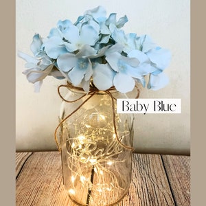 May include: A mason jar with a string of fairy lights inside and a bouquet of light blue hydrangeas on top. The jar is labeled "Baby Blue".