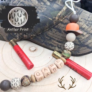 May include: A camouflage baseball cap with a decorative charm featuring a miniature orange baseball cap, wooden beads, and red shotgun shells. The charm includes the name "GABE" spelled out on wooden blocks. The image also includes the text "Antler Print".