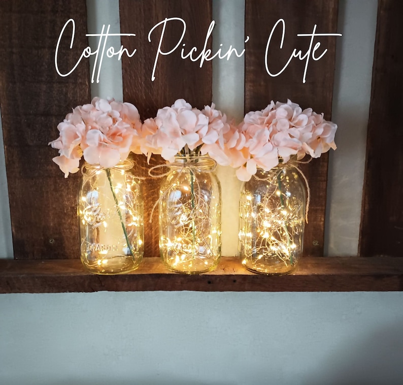May include: Three mason jars filled with pink flowers and fairy lights. The jars are sitting on a wooden shelf. The text "Cotton Pickin' Cute" is written above the jars.