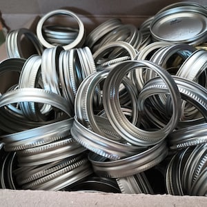 May include: A close-up of a cardboard box filled with silver metal jar lids.
