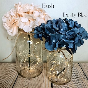 May include: Two mason jars with string lights inside, decorated with blush and dusty blue hydrangea flowers. The jars are sitting on a wooden surface.