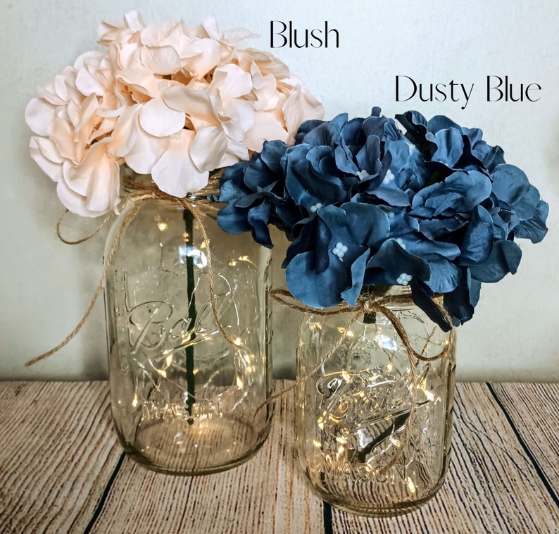 May include: Two mason jars with string lights inside, decorated with blush and dusty blue hydrangea flowers. The jars are sitting on a wooden surface.
