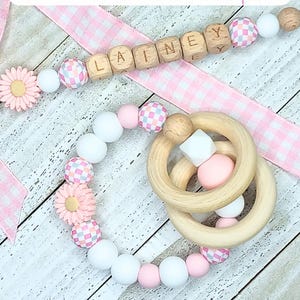 May include: A pink and white baby teether with a wooden ring and a wooden bead necklace with the name "Lainey" spelled out. The necklace and teether are on a pink and white gingham ribbon. The text "Cotton Pickin' Cute Baby" is at the bottom of the image.