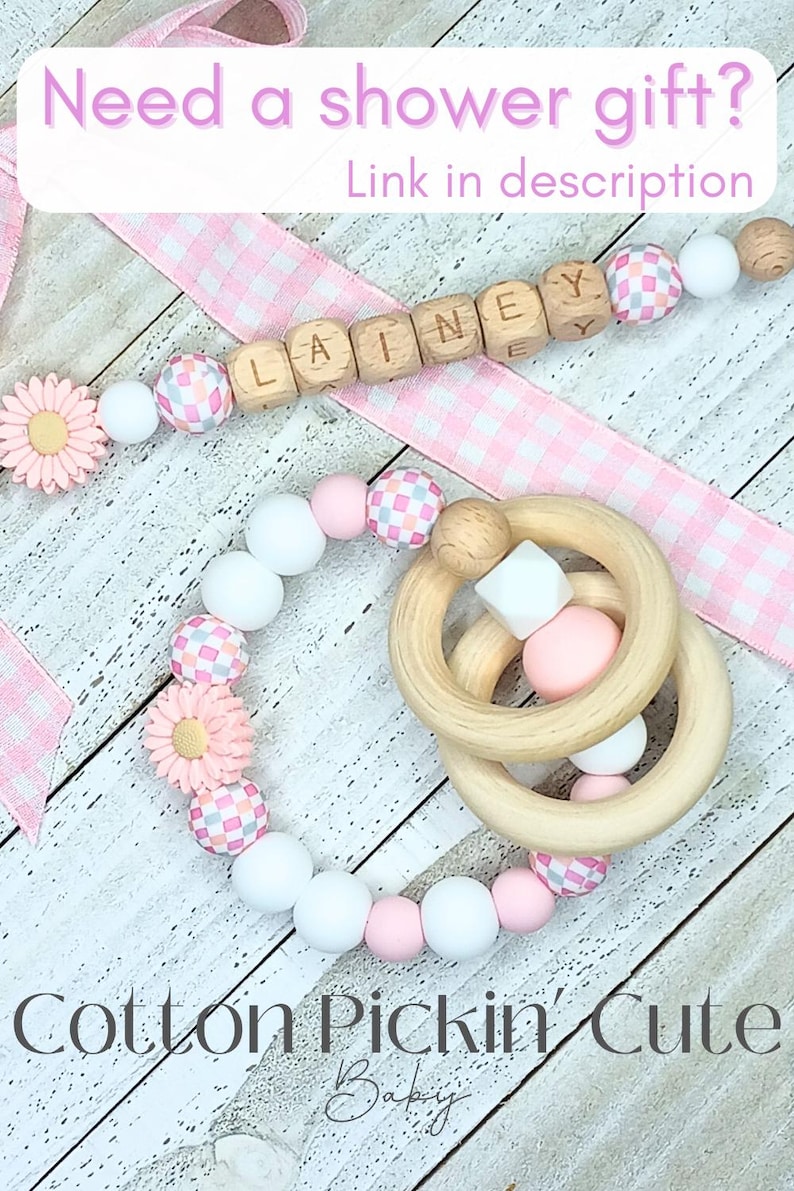 May include: A pink and white baby teether with a wooden ring and a wooden bead necklace with the name "Lainey" spelled out. The necklace and teether are on a pink and white gingham ribbon. The text "Cotton Pickin' Cute Baby" is at the bottom of the image.