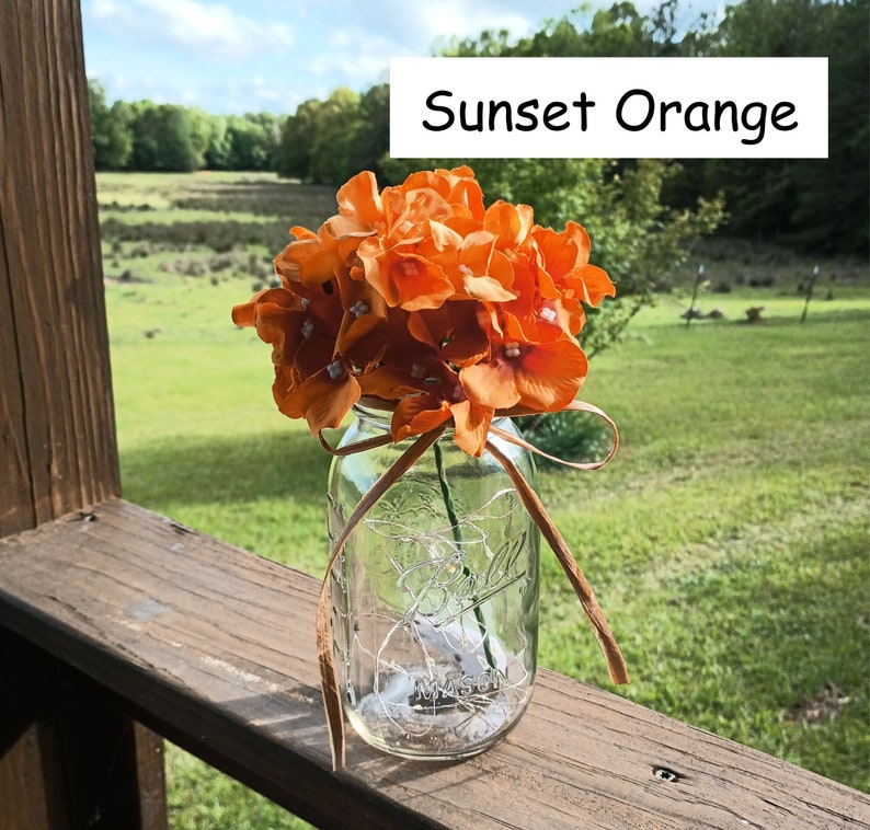 May include: A mason jar filled with a bouquet of orange silk flowers, tied with a brown ribbon. The jar is sitting on a wooden surface. The text "Sunset Orange" is visible in the image.