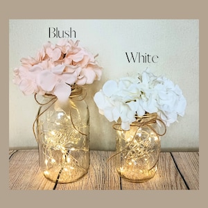 May include: Two mason jars with string lights inside, decorated with blush and white hydrangea flowers. The jars are labeled "Blush" and "White".