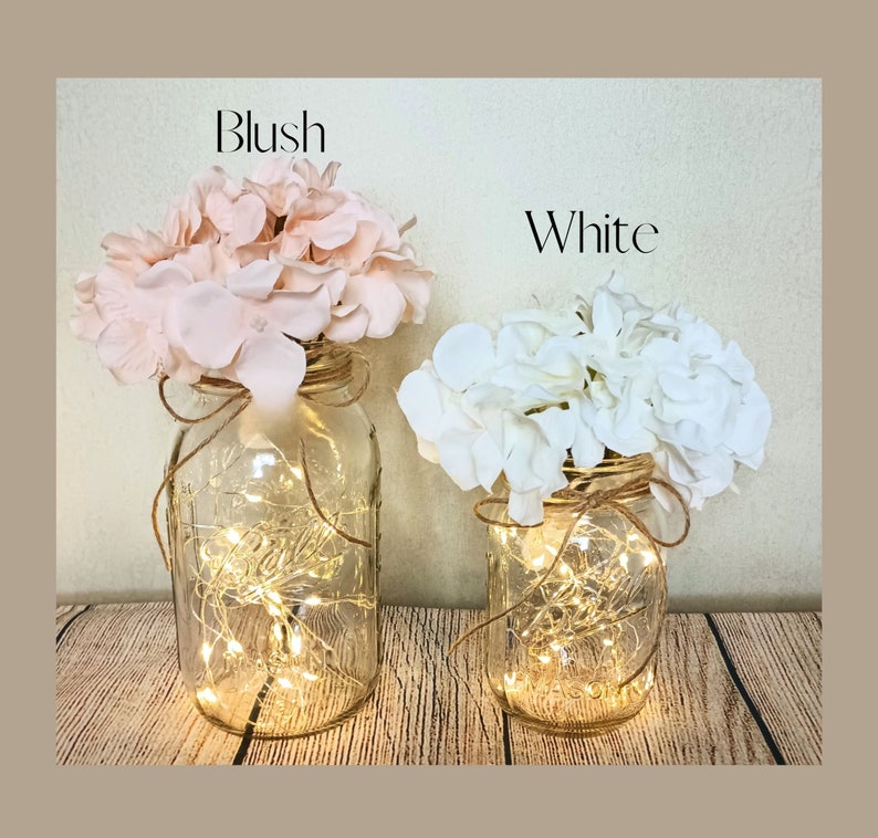 May include: Two mason jars with string lights inside, decorated with blush and white hydrangea flowers. The jars are labeled "Blush" and "White".