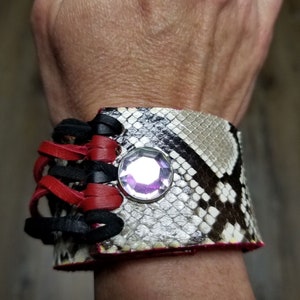 May include: A black and red leather bracelet with a snakeskin pattern and a clear rhinestone. The bracelet has a lace-up design.